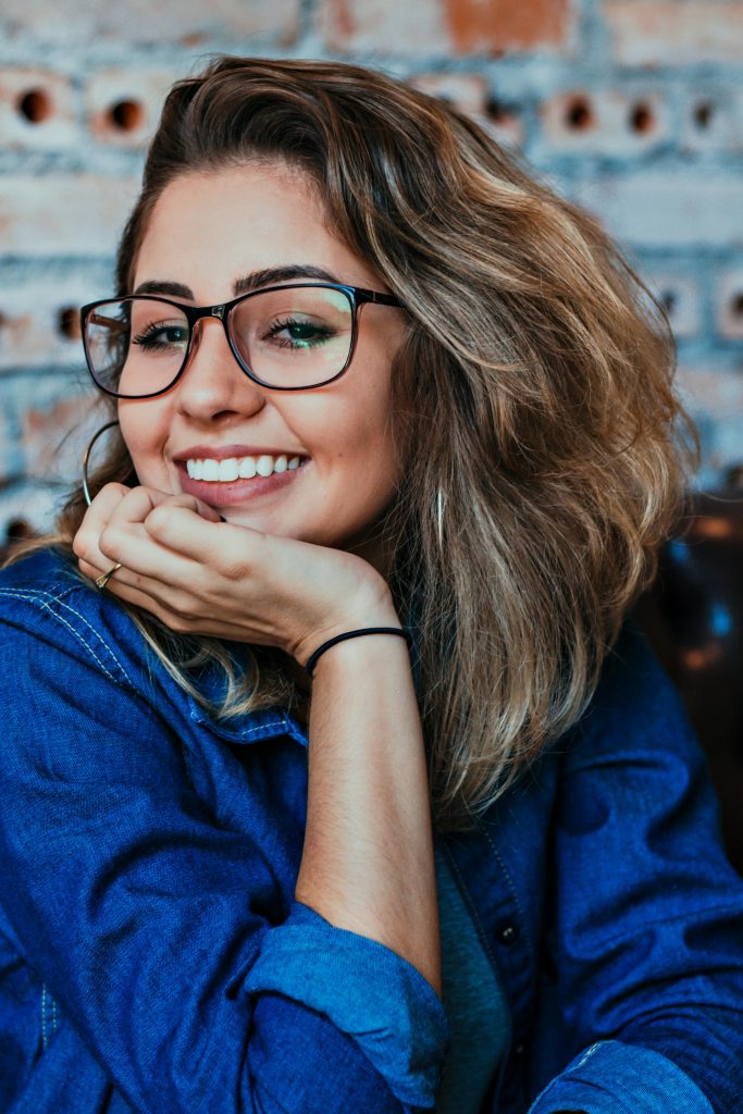 A young woman wearing glasses and denim, smiling in an indoor setting.